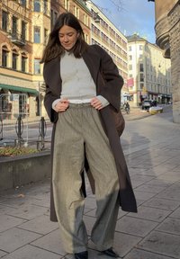 Manteau en laine brun foncé sur un pull à boutons gris clair, associé à un pantalon large rayé gris et des bottines noires.