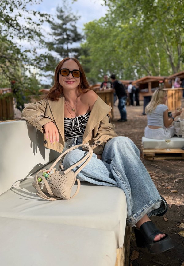 Joven mujer con gafas de sol, chaqueta beige, top a rayas y jeans anchos sentada al aire libre en un banco blanco con bolso beige, árboles y personas en el fondo.