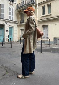 Cappotto trench beige, jeans a gamba larga blu scuro e scarpe color crema con accenti verdi. Cappellino arancione e zaino marrone, in piedi su una strada di città.