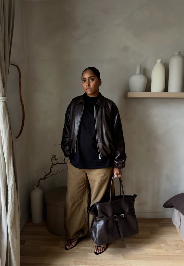 Osoba stojaca vo vnútri, oblečená v čiernej koženej bunde a hnedom nohaviciach, drží veľkú čiernu koženú tašku pri vázy na poličke na stene.