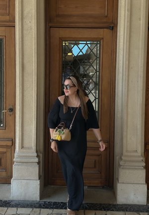 Largo vestido negro sin hombros, zapatos planos en tono taupe, bolso de mimbre con acentos coloridos, enmarcado por puertas de madera y arquitectura de piedra.