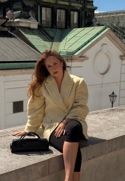 Joven mujer con abrigo beige y pantalones negros sentada en un borde de piedra con bolso negro, techo de edificio histórico en el fondo bajo cielo despejado.