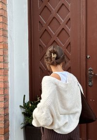 Pull en grosse maille crème avec une coupe ample, tricot texturé. Sac à main marron sur le côté, plante verte dans un panier à proximité, porte marron en arrière-plan.