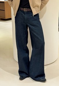 Szerokie ciemnoniebieskie dżinsy o teksturowanym wykończeniu. Wyposażone w zamek błyskawiczny i szlufki na pasek. Połączone z jasnobrązową kurtką i ciemnym obuwiem.