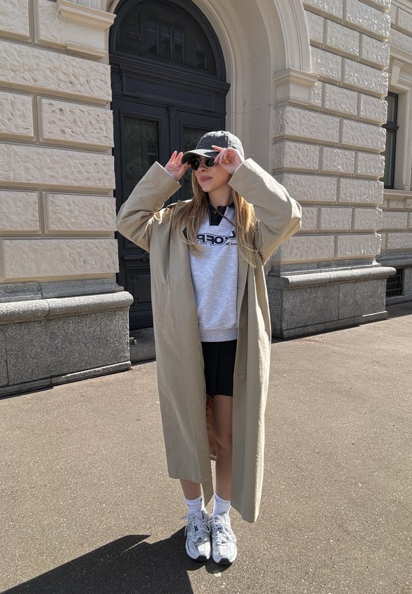 Beiger Oversize-Mantel, grauer Hoodie mit Logo, schwarze Shorts, weiße Socken und graue Sneaker. Zubehör umfasst Sonnenbrille und eine Mütze.