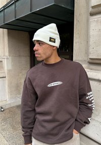 Νέος άνδρας με φακίδες που φοράει λευκό σκουφάκι και καφέ φούτερ με λευκό κείμενο, στέκεται δίπλα σε τοίχο από πέτρα.