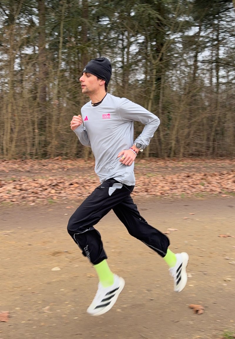 Δρομέας που φοράει γκρι μπλούζα με μακρύ μανίκι, μαύρο παντελόνι, νέον πράσινες κάλτσες και άσπρα παπούτσια, τρέχοντας σε μονοπάτι δάσους το φθινόπωρο.