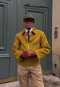 Άνδρας που φοράει μουσταρδί μπουφάν, χακί παντελόνι, καφέ γάντια, ελιά καπέλο και μεγάλα γυαλιά ηλίου στέκεται μπροστά από γκρι πόρτες.