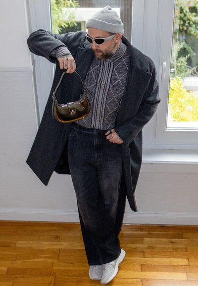 Persona con un abrigo largo gris sobre un suéter estampado, jeans oscuros, zapatos blancos y sosteniendo un pequeño bolso brillante.