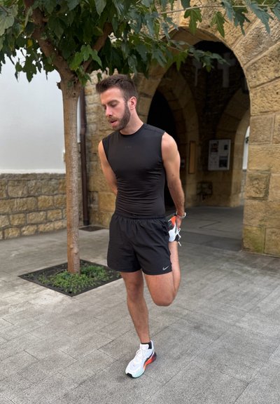 Top negro sin mangas con logo, pantalones cortos negros, zapatillas deportivas blancas y de colores. Estirando la pierna mientras está de pie cerca de un árbol y un muro de piedra.