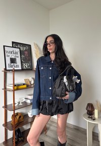 Denim jakna s srebrnimi gumbi in modrimi poudarki, v kombinaciji s črno plisirano krilo. Vključuje teksturirano črno torbico s dekorativnimi elementi.