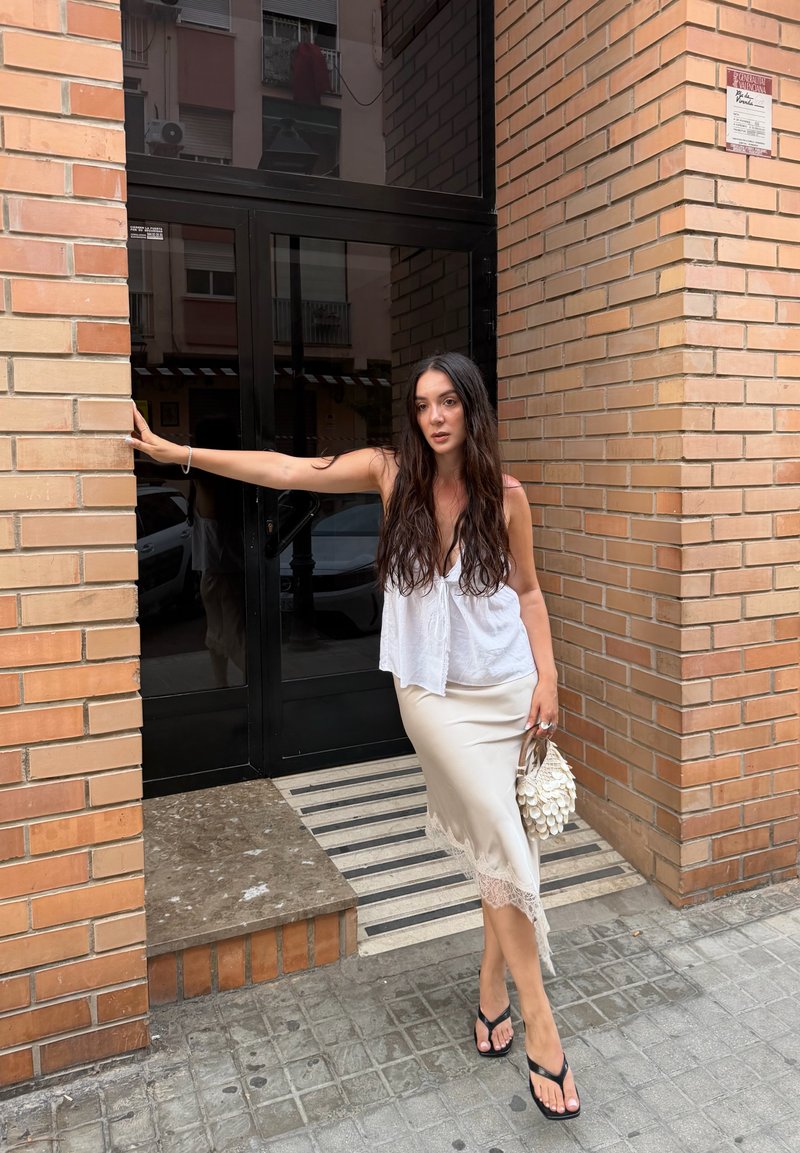 Femme aux longs cheveux foncés portant un haut blanc sans manches, une jupe beige bordée de dentelle, et des sandales noires, debout près de l'entrée d'un bâtiment en briques, tenant un petit sac.