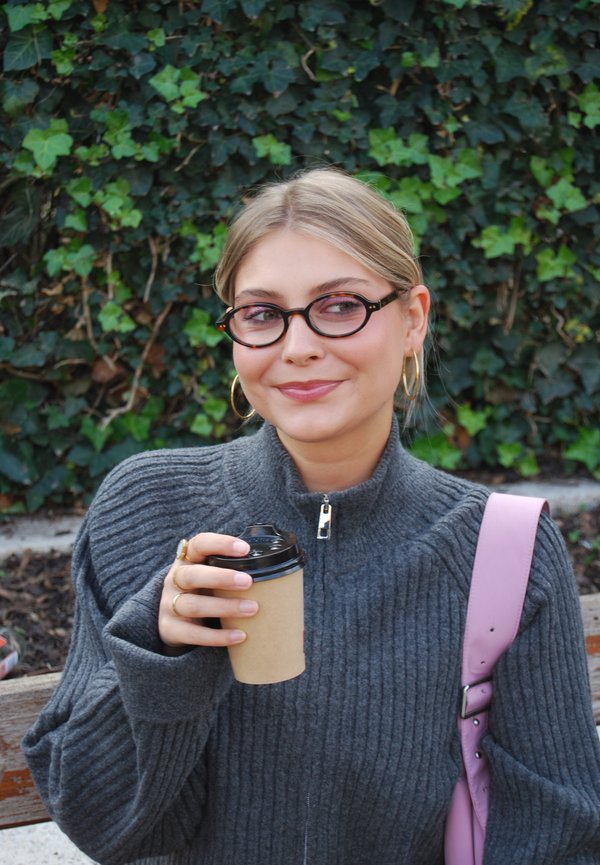 Pull en ribbed gris avec col haut, tenant une tasse en papier marron avec un couvercle noir et portant des lunettes rondes avec une monture noire et verte.