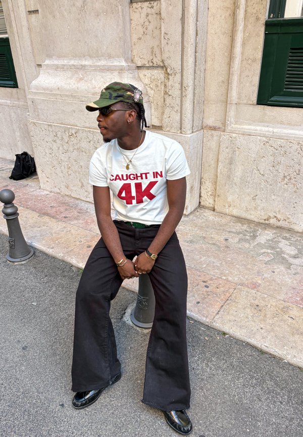 Uomo seduto su un breve paletto stradale, indossa una t-shirt bianca "Caught in 4K", pantaloni neri, cappellino mimetico, occhiali da sole e scarpe nere.