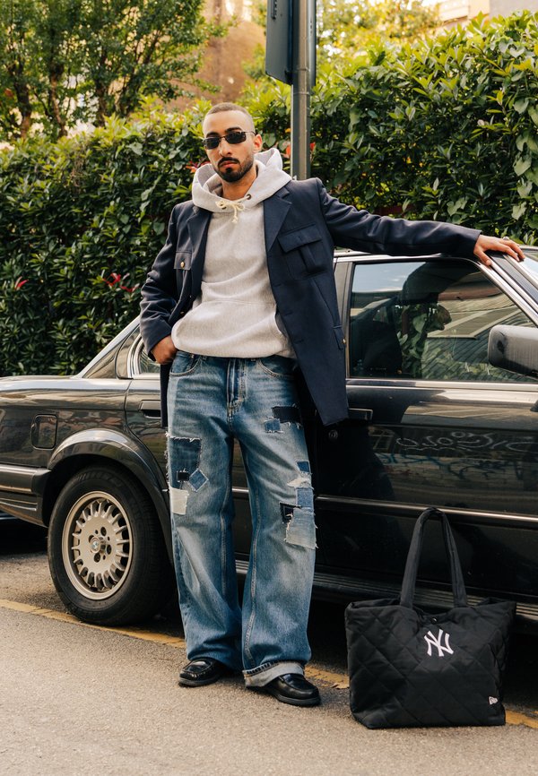 Hombre con gafas de sol que lleva un abrigo azul marino, una sudadera gris con capucha y vaqueros de retales, está junto a un coche negro con una bolsa acolchada negra de los New York Yankees en el suelo.