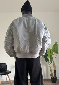 Padded bomber jacket in light gray, featuring ribbed cuffs and hem, with shoulder and side gathers. Worn with black wide-leg pants.
