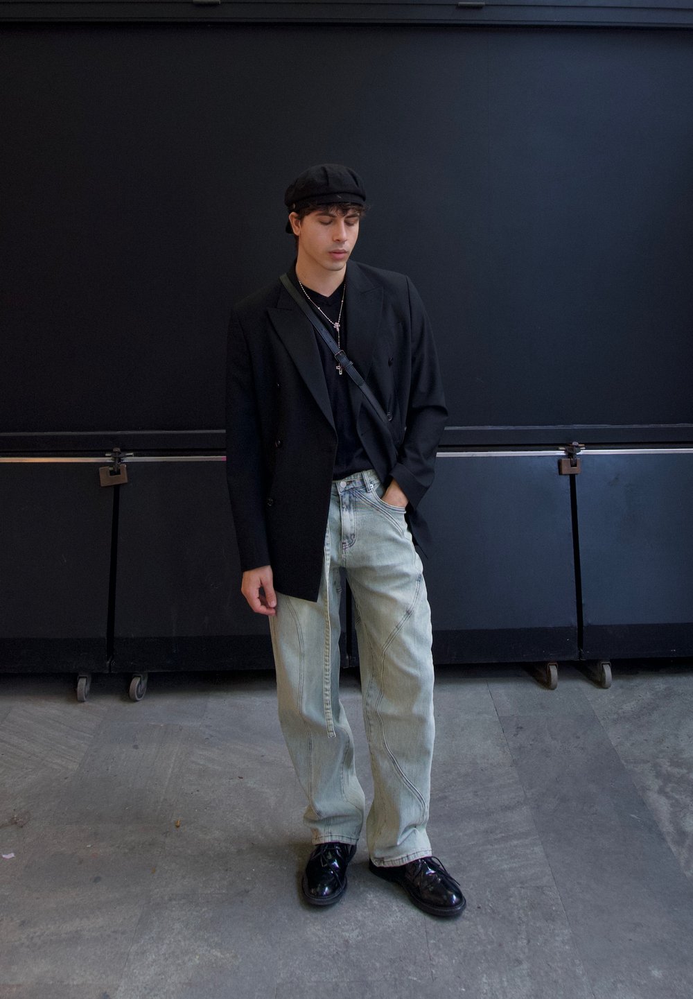 Blazer nero doppiopetto sopra una t-shirt nera, jeans baggy azzurri chiari e scarpe nere lucide. Si indossa un berretto nero e collane a strati.