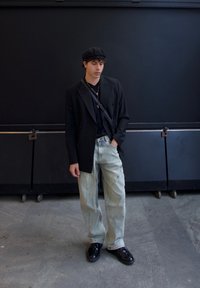 Blazer nero doppiopetto sopra una t-shirt nera, jeans baggy azzurri chiari e scarpe nere lucide. Si indossa un berretto nero e collane a strati.