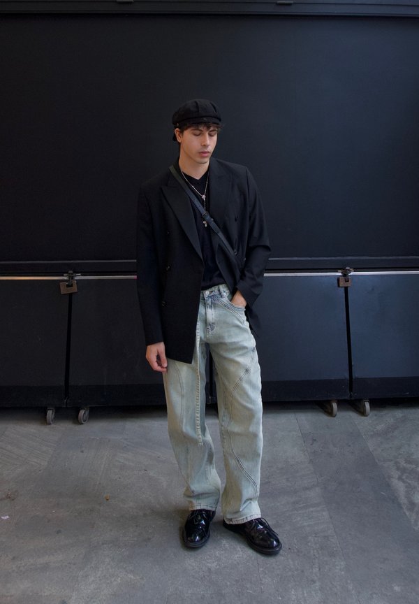 Blazer nero doppiopetto sopra una t-shirt nera, jeans baggy azzurri chiari e scarpe nere lucide. Si indossa un berretto nero e collane a strati.