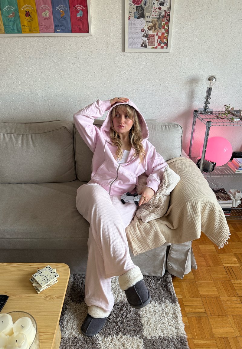 Femme en tenue décontractée rose et chaussons assise sur un canapé gris tenant une manette de jeu dans un salon cosy avec des œuvres murales et une table d'appoint.