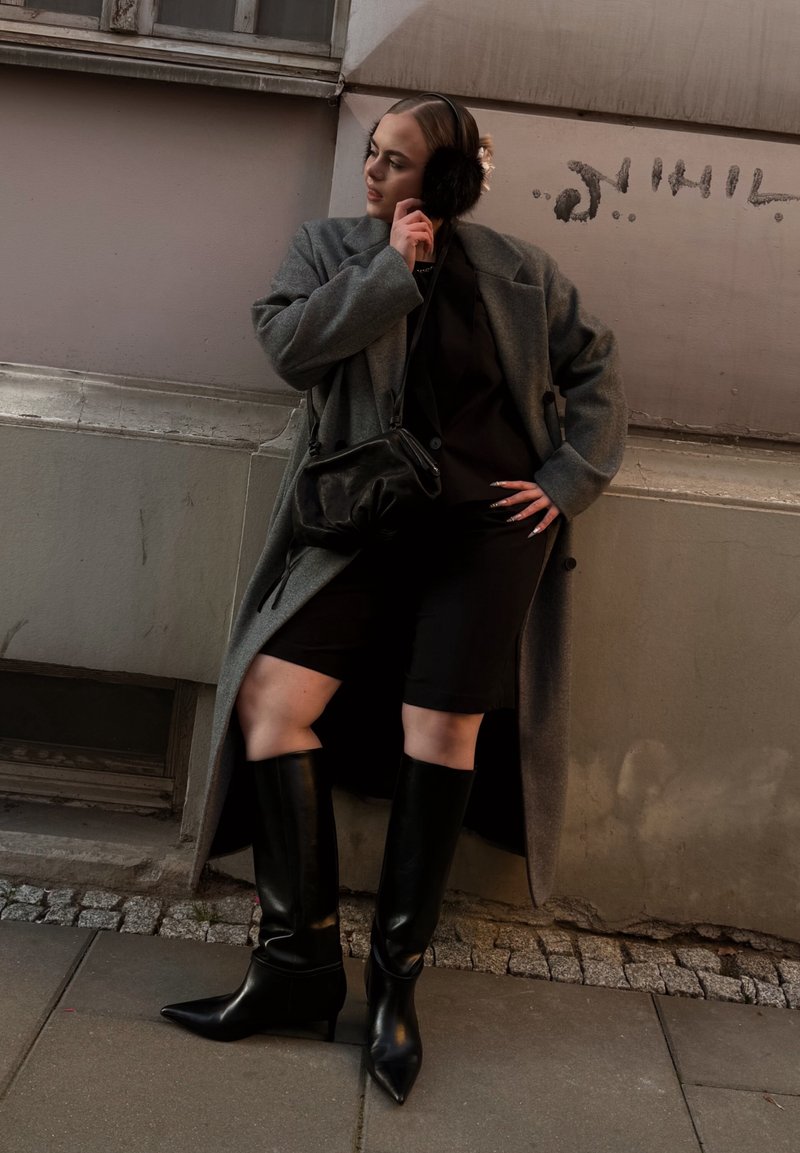 Gray wool coat, black fitted dress and knee-high black heeled boots. Features faux fur ear muffs and a small black shoulder bag.