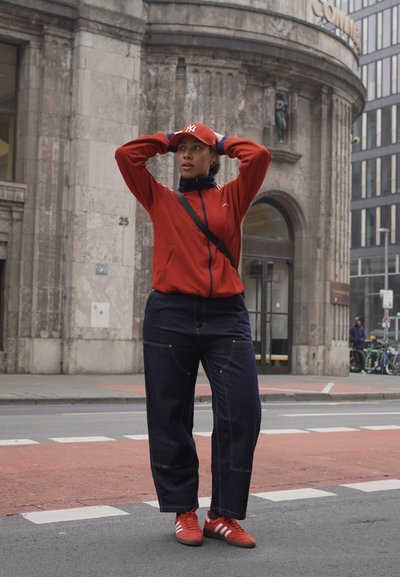 Chaqueta roja con cremallera, pantalones vaqueros de pierna ancha en denim oscuro, zapatillas rojas con rayas blancas y una gorra roja. Fondo urbano con detalles de edificios de piedra.