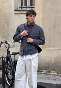 Hall hõlmikust jope kõrge kraega, nelja eesmise tasku ja nööbidetailidega, kombineeritud valgete linaste pükste ja õlakotiga.