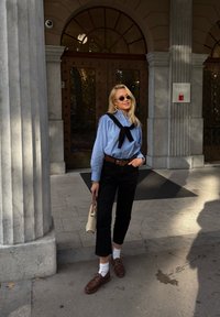 Giovane donna con occhiali da sole si trova all'esterno di un edificio con grandi porte in legno, indossando una camicia blu, pantaloni neri e scarpe marroni, mentre tiene una borsa.