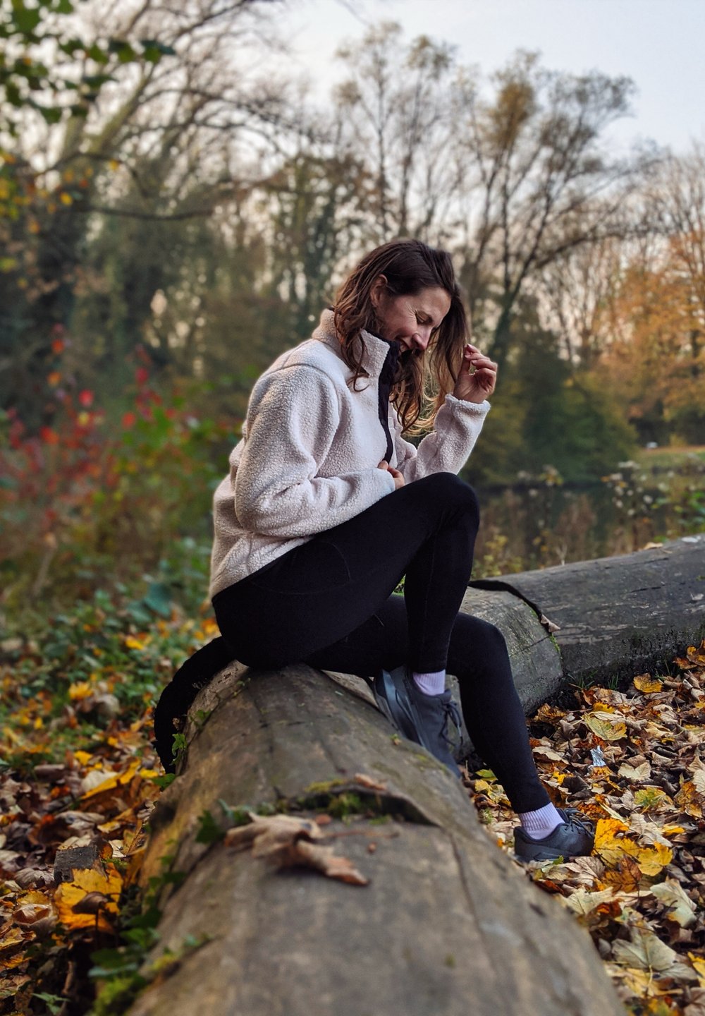 Casaco de fleece bege claro com fecho, leggings pretas e sapatos cinzentos usados por uma pessoa sentada numa tronco rodeada de folhas caídas numa área florestal.