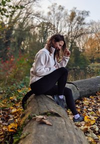 Casaco de fleece bege claro com fecho, leggings pretas e sapatos cinzentos usados por uma pessoa sentada numa tronco rodeada de folhas caídas numa área florestal.
