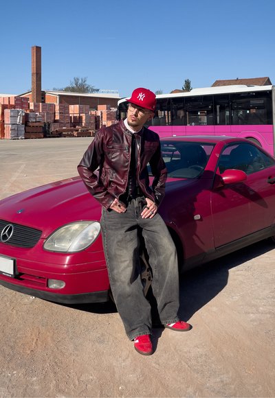Chaqueta bomber de cuero, vaqueros grises anchos, gorra roja y zapatos rojos. Mercedes coupe rosa al fondo, con un entorno industrial y cielo azul.