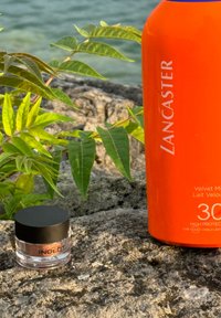En orange solskyddsflaska märkt "Lancaster Velvet Milk SPF 30" bredvid en genomskinlig behållare med Inglot shimmerpulver med svart lock på en sten.