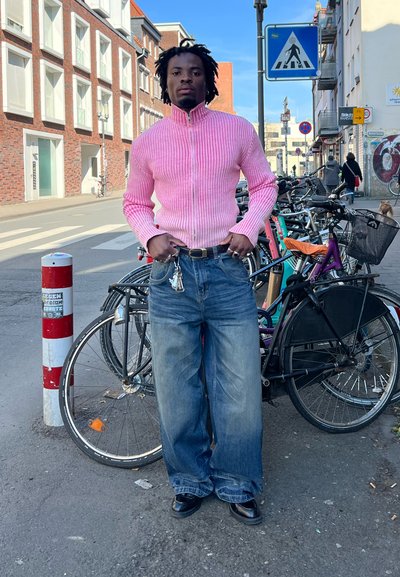 Suéter de cremallera acanalado rosa, vaqueros anchos azul claro, zapatos negros. En el fondo hay bicicletas y una señal de tráfico.