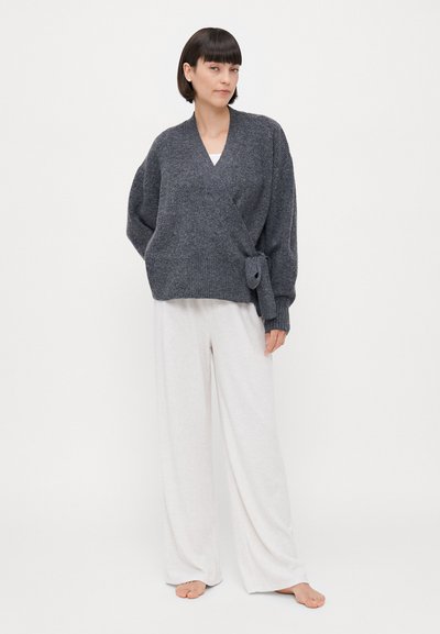 Suéter envolvente de punto gris con un detalle atado en la cintura, combinado con pantalones de pierna ancha de color claro. Textura suave, ajuste relajado.