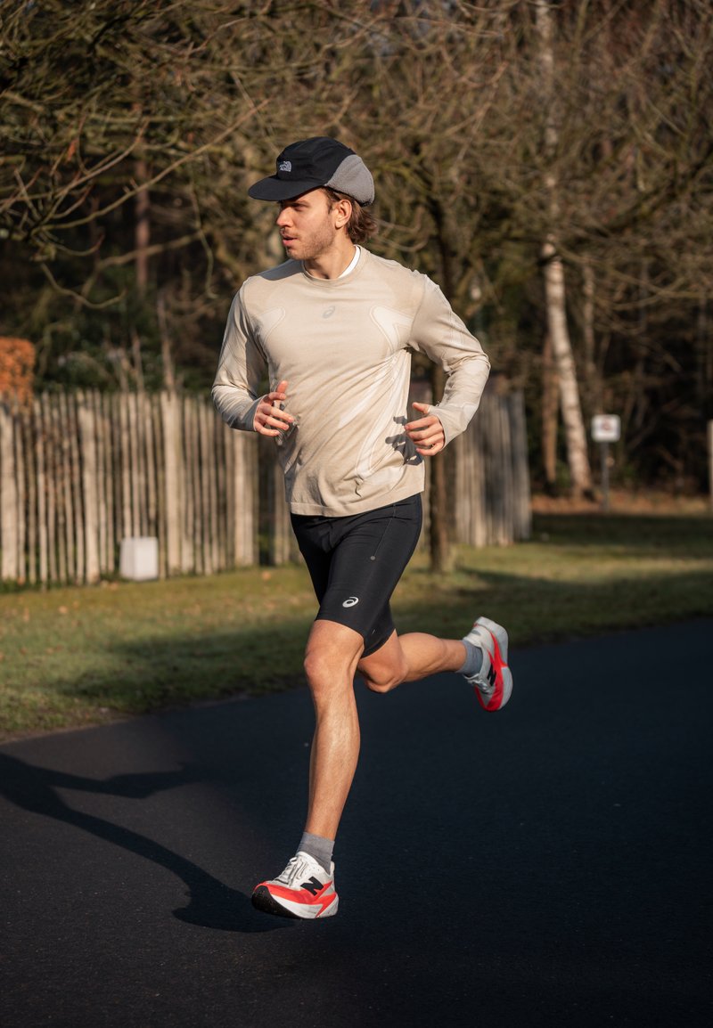 Vyras su kepure, dėvintis smėlio spalvos ilgomis rankovėmis marškinius ir juodus šortus, bėgioja takeliu parke, kur nuogi medžiai ir medinės tvoros.