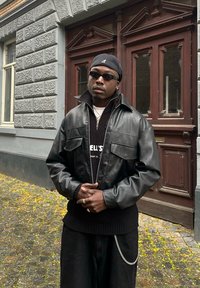 Black leather jacket with a wide collar and two front pockets, worn with a black knitted sweater and dark trousers featuring a silver chain.