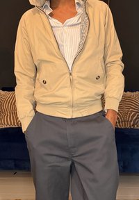 Personne portant une veste beige à fermeture éclair sur une chemise rayée, les mains dans les poches d’un pantalon gris, debout à l’intérieur devant des coussins à motif.