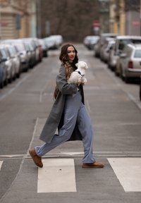 Femme traversant un passage pour piétons en ville en tenant un petit chien blanc, portant un manteau gris, un pantalon bleu et des chaussures marron.