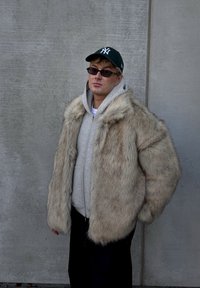 Nepbontjas in lichtbeige met een pluizige textuur, gedragen over een grijze hoodie. Zwarte broek en een groene pet maken de look compleet.