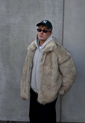 Abrigo de piel sintética en beige claro con una textura esponjosa, llevado sobre una sudadera con capucha gris. Pantalones negros y una gorra verde completan el conjunto.