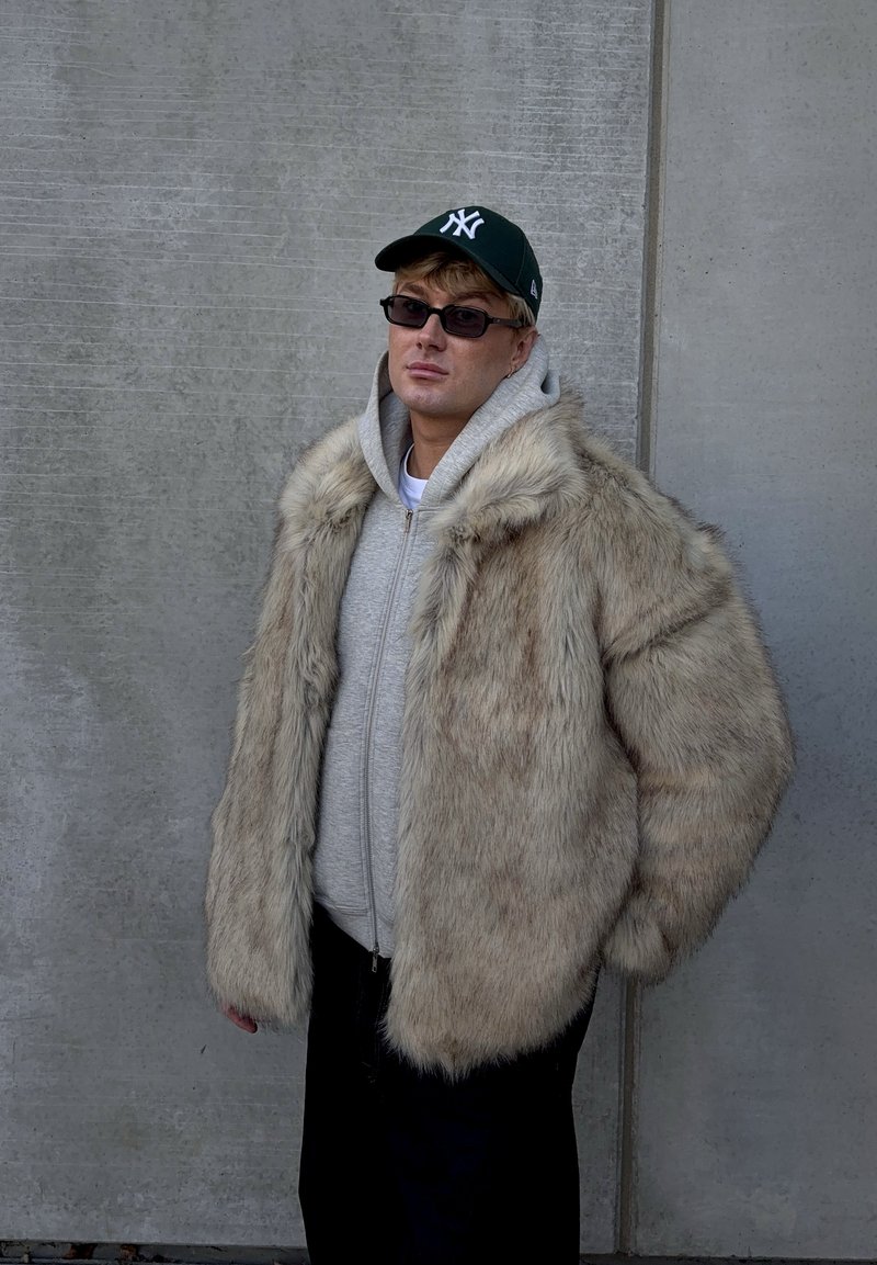 Cappotto in pelliccia sintetica color beige chiaro con una texture soffice, indossato sopra una felpa grigia con cappuccio. Completa il look un paio di pantaloni neri e un berretto verde.