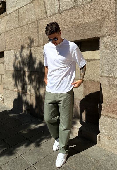 Camiseta blanca, pantalones verde oliva y zapatillas blancas. Tejido suave y corte relajado. De pie sobre un pavimento de piedra con una pared texturizada atrás.