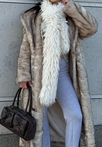 Long manteau en fausse fourrure beige avec un large col, écharpe blanche texturée et shaggy, haut gris ajusté, pantalon gris clair et sac à main noir avec deux poches.