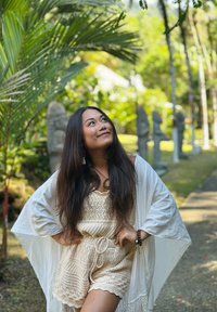 Γυναίκα με κρεμ πλεκτό σύνολο και λευκή εσάρπα στέκεται με τα χέρια στη μέση, κοιτώντας προς τα πάνω, περιτριγυρισμένη από τροπικά φυτά και πέτρινα αγάλματα.