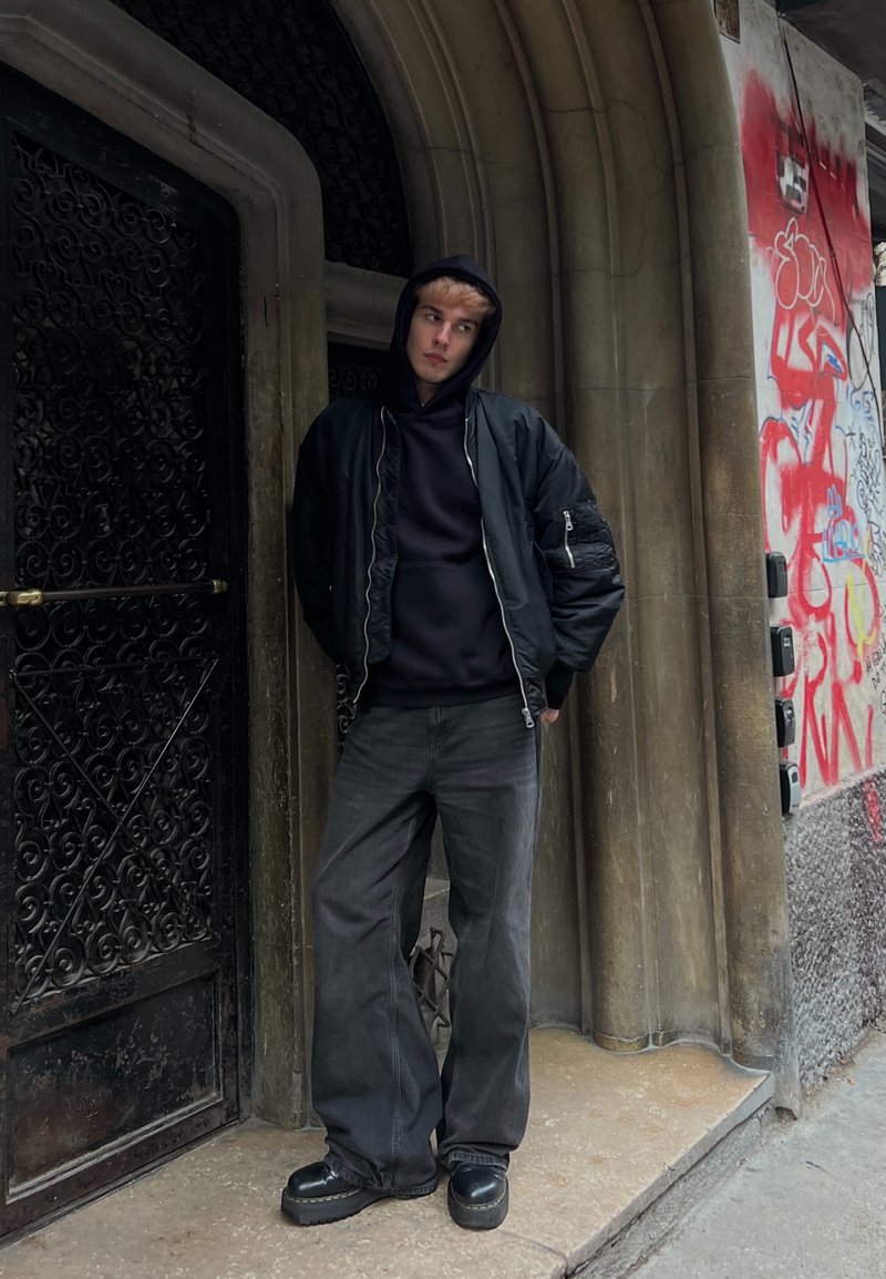 Jeune personne portant un sweat à capuche noir, une grande veste noire, un jean noir large et des chaussures à plateforme, debout devant une porte ornée et un mur de graffiti.