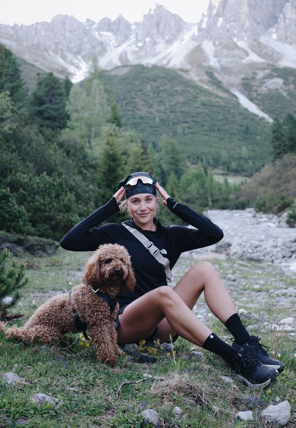 Mladá žena v čiernom outdoorovom oblečení sedí na tráve s kučeravým hnedým psom, v pozadí hory a les.
