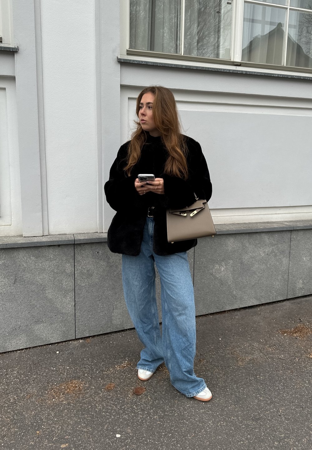 Μαύρο τεχνητό γούνινο μπουφάν, ανοιχτό μπλε φαρδιά τζιν παντελόνια και λευκά παπούτσια. Κρατάει ένα smartphone και μία καφέ τσάντα με χρυσές λεπτομέρειες.