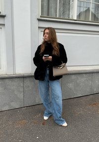 Μαύρο τεχνητό γούνινο μπουφάν, ανοιχτό μπλε φαρδιά τζιν παντελόνια και λευκά παπούτσια. Κρατάει ένα smartphone και μία καφέ τσάντα με χρυσές λεπτομέρειες.