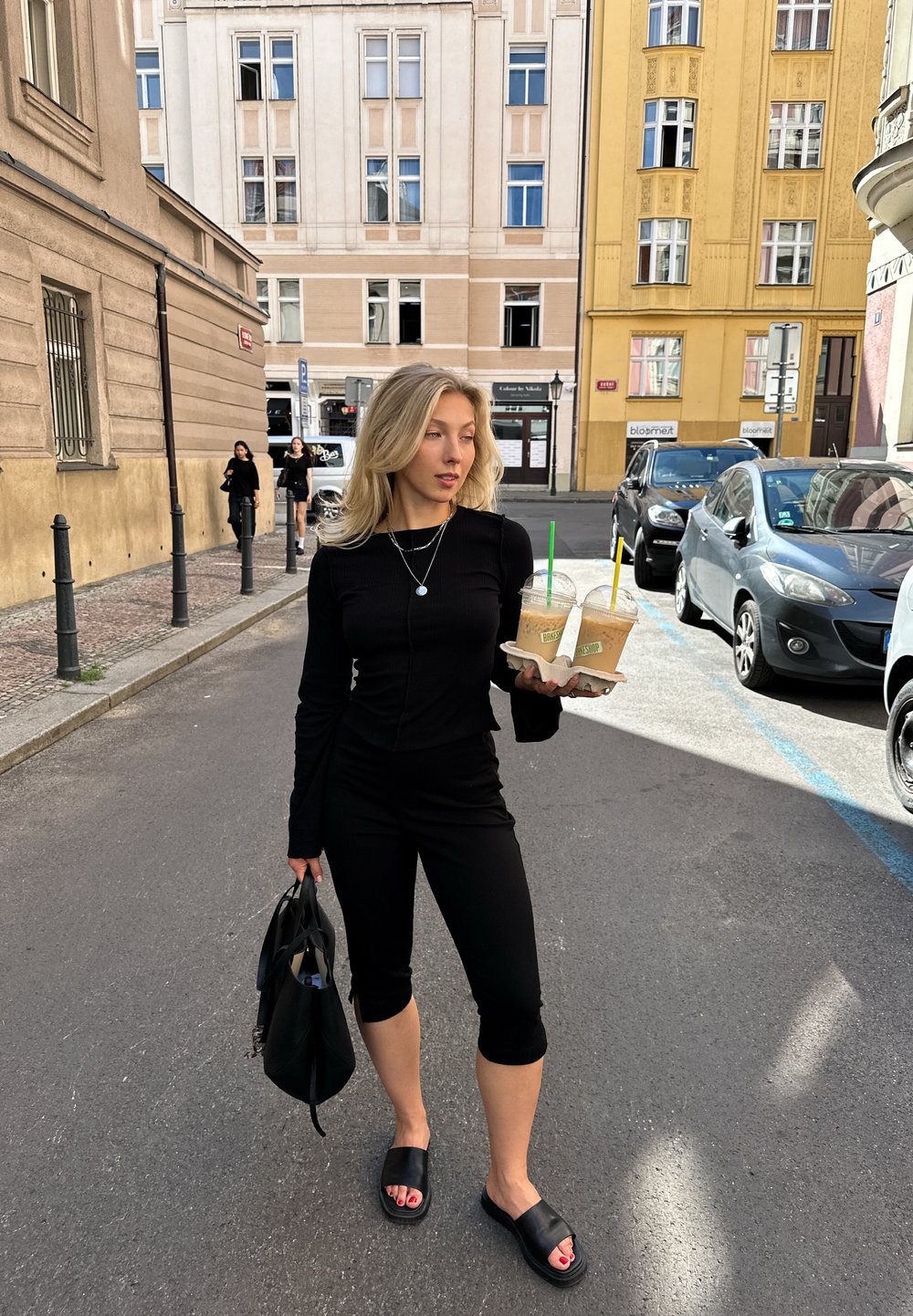 Čierny priliehavý top s dlhým rukávom a legíny, drží dva ľadové nápoje, s textúrovanou čiernou taškou a čiernymi šľapkami. Pozadie ulice.