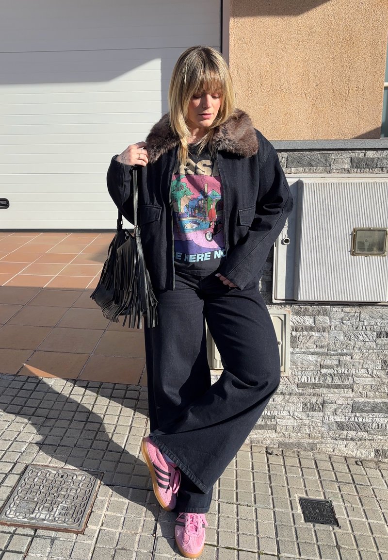 Nő fekete szőrmés galléros kabátban, mintás pólóban, széles fekete nadrágban, rózsaszín Adidas tornacipőben, fekete rojtos táskát tartva, a járdán áll.